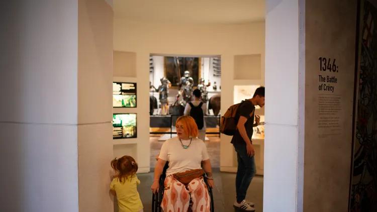 a visitor in a wheelchair navigates a gap in a gallery comfortably while passing a small child who leans against the wall