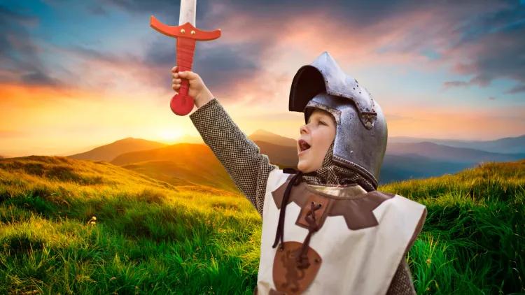 Child dressed as a knight holding a red handled sword and pointing it towards the sky with a green hilly background as sunset
