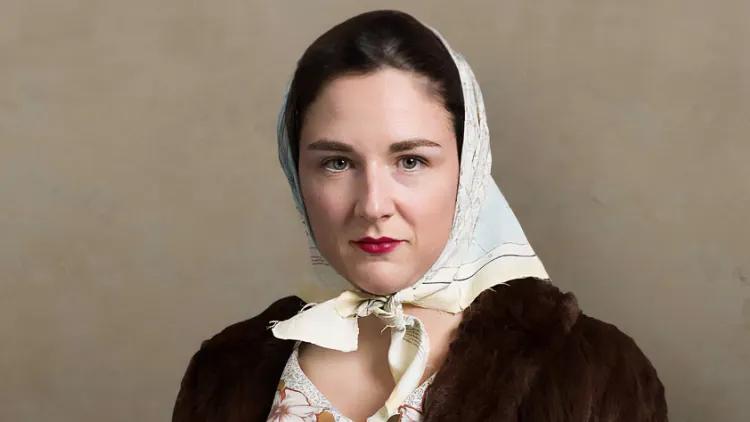 A young female SOE agent with a steely gaze poses in a fur coat and carries a sub-machine gun