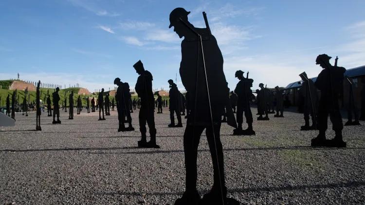 Life-sized soldier silhouettes positioned on a stony ground, heads bowed, evoking remembrance against a bright sky backdrop