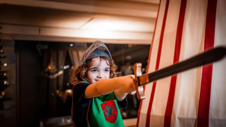 A child dressed in medieval-style costume wearing a metal-look helmet and holding a long toy sword outward, standing beside a canvas structure with red vertical stripes.