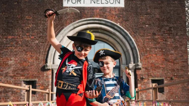 Two young children dressed as pirates pose with raised swords on a bridge, with a large brick entrance behind them displaying the text “FORT NELSON”