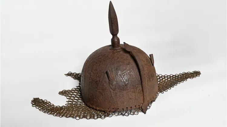 Historical metal helmet featuring a rounded top with a pointed finial and a chainmail neck guard extending outward on both sides