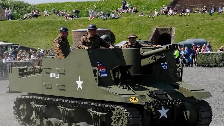 Olive-green self-propelled artillery vehicle with white star markings, moving on gravel with people aboard and a crowd behind