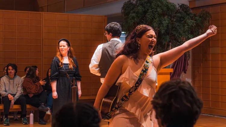 Performer in light dress with guitar strap raises arm while singing on small stage; other cast members and seated audience visible
