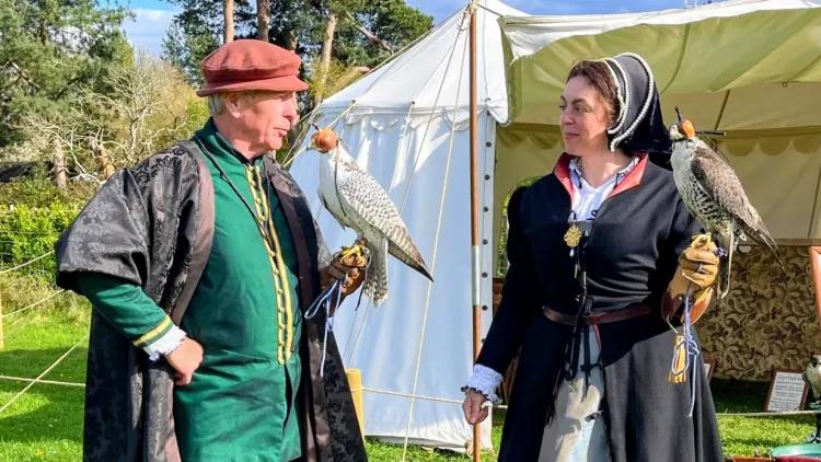 Falconry display outdoors: two costumed people with hooded falcons stand on grass near a white tent and woodland