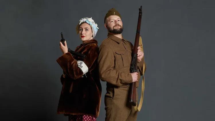 A Second World War female SOE operative and soldier standing back to back looking forward.  The SOE is in civilian dress with a fur coat and head scarf. The soldier is in uniform. Both are holding firearms