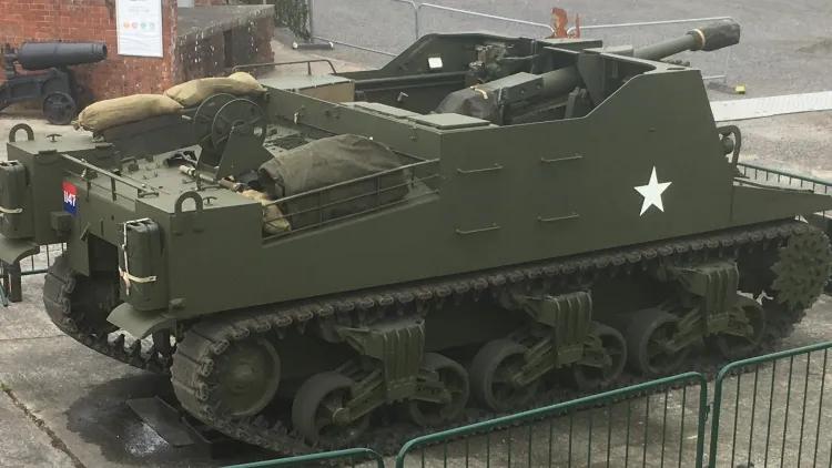 Sexton tracked gun carrier with white star marking, stationed by seaside fort with barriers and misty background