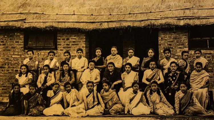 Sepia-toned group of women in saris and uniforms, seated and standing in three rows in front of a brick structure