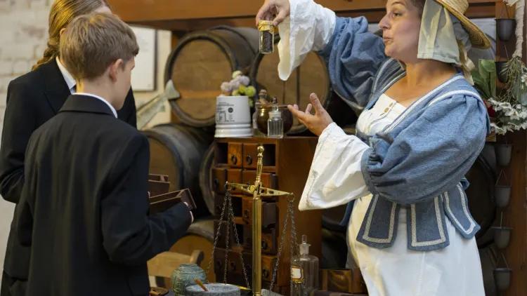 17th-century herbal women displays medicine jar to two school pupils