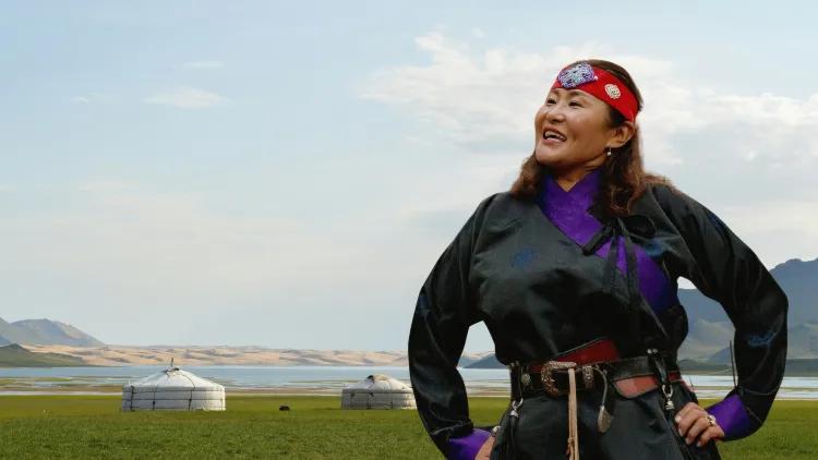 Figure in black attire with purple trim overlooking open plains, yurts and a calm lake under a bright sky