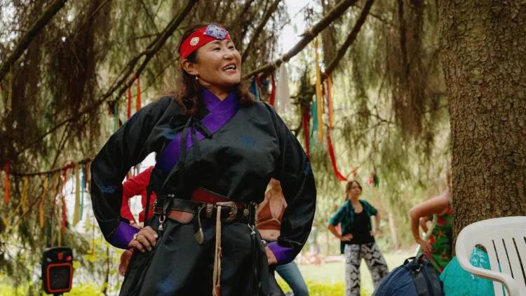Person in black and purple ceremonial clothing with a red headband stands beneath trees decorated with colourful ribbons