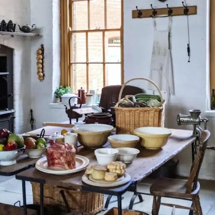 19th century kitchen with 2 large windows a stove, table with bowls and food items and 2 chairs