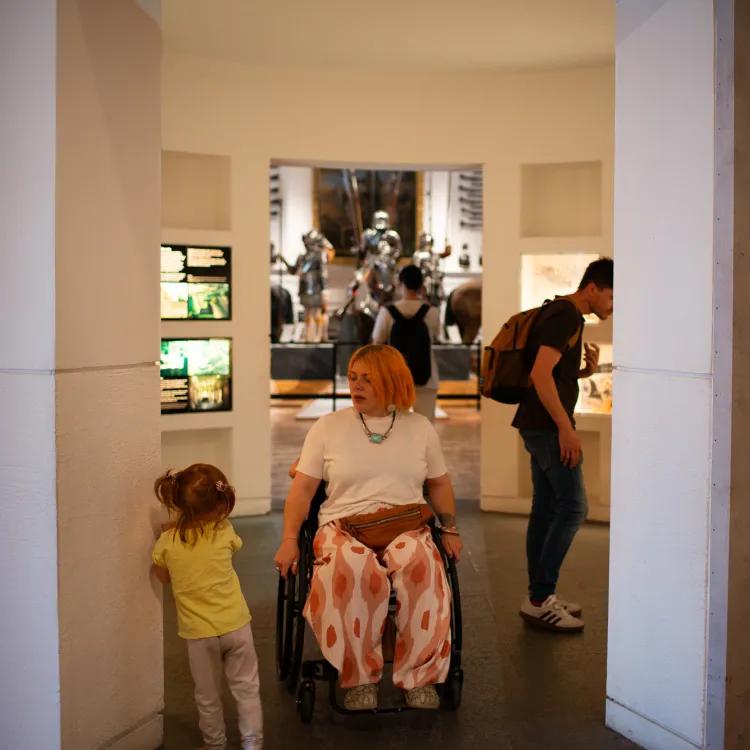 a visitor in a wheelchair navigates a gap in a gallery comfortably while passing a small child who leans against the wall