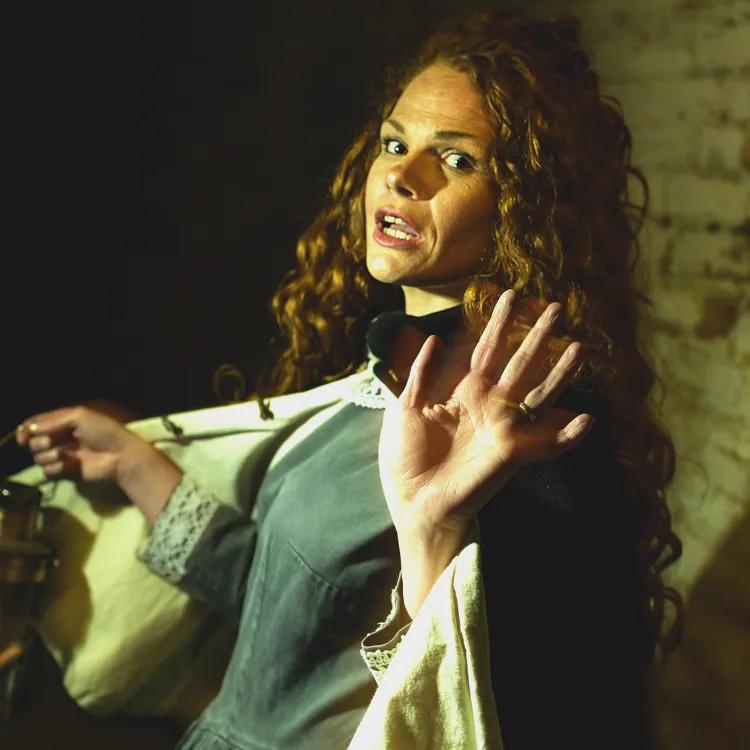 woman with red curly hair dressed in green dress and cloak and holding a lamp raises a beckoning hand 