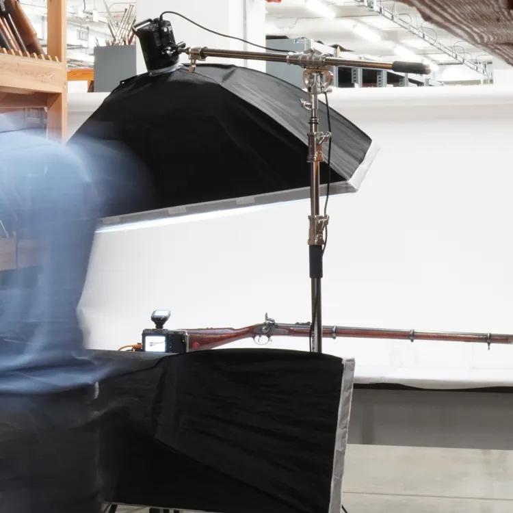 Photographer looking at image on a laptop with firearm on a table with photography lights and equipment around it.