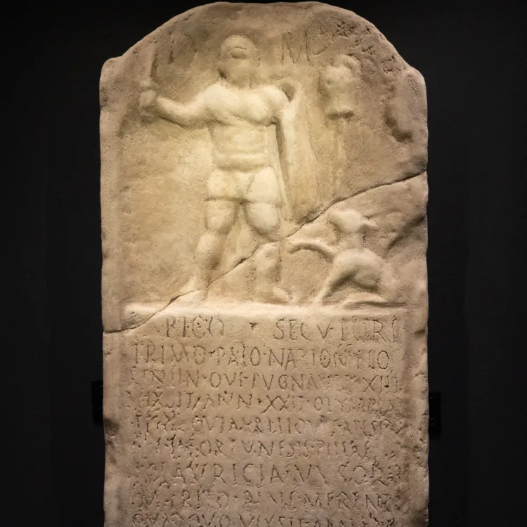 A sandy coloured stone tablet depicting a gladiator and his dog, with his life story inscribed below