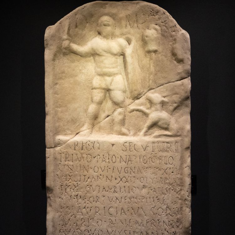 A sandy coloured stone tablet depicting a gladiator and his dog, with his life story inscribed below