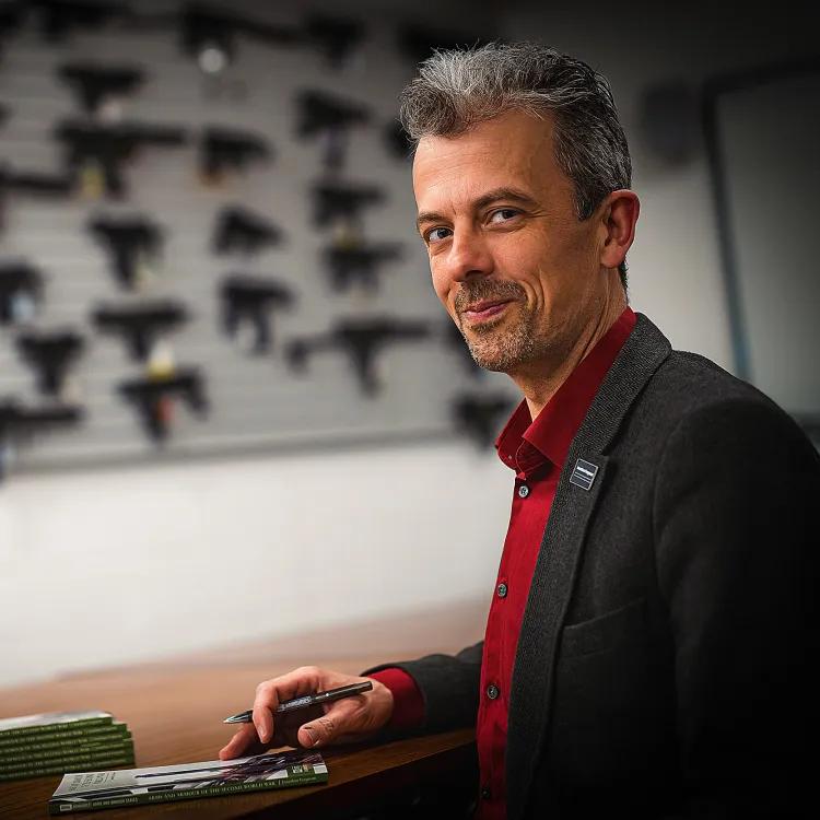 Jonathan Ferguson sitting at a table signing books with guns displayed in the background