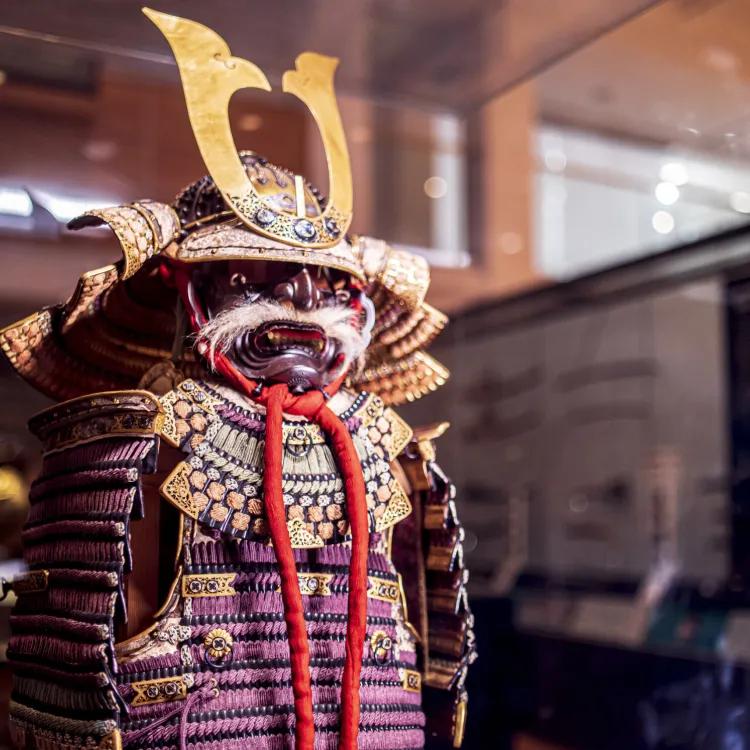 a purple and gold Samurai armour with a fearsome mask in the form of a demon with white whiskers stands guard in the gallery