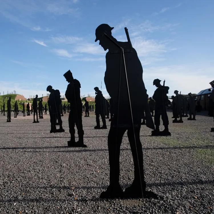 Life-sized soldier silhouettes positioned on a stony ground, heads bowed, evoking remembrance against a bright sky backdrop