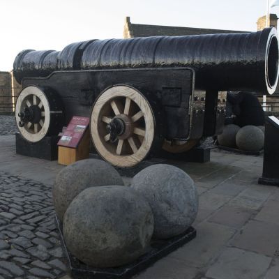 A bombard from the 15th century. An enormous barrel made from black metal mounted on a four wheeled cart. Three huge stone balls are placed on the floor in front on it. 