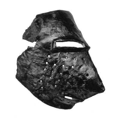 Right side of a corroded fragment of a metal visor with narrow eye and mouth slit, multiple holes on cheek and a pointed snout