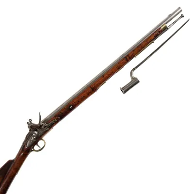 A long barrel carbine with handrail carved buttstock, finished in wood, steel and slight brass trim. A steel bayonet lies to the side.