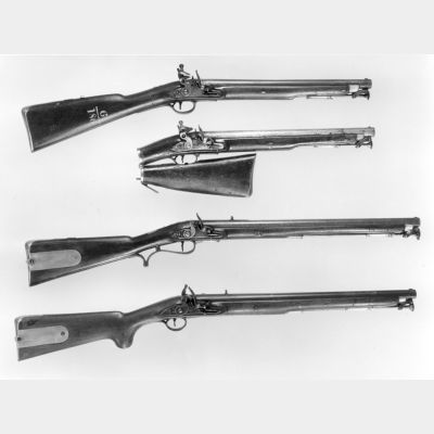 A black and white image of four short barrelled carbines in wood and steel, with brass trim