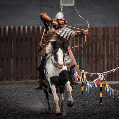 An actor on horseback in Mongolian armour draws and aims a bow at a target off screen