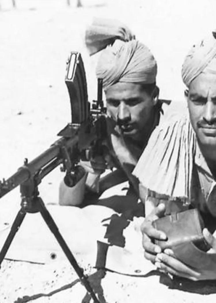 Two prone Indian army soldiers in turbans operate a tripod-mounted Berthier light machine gun, one aiming while the other manages ammunition