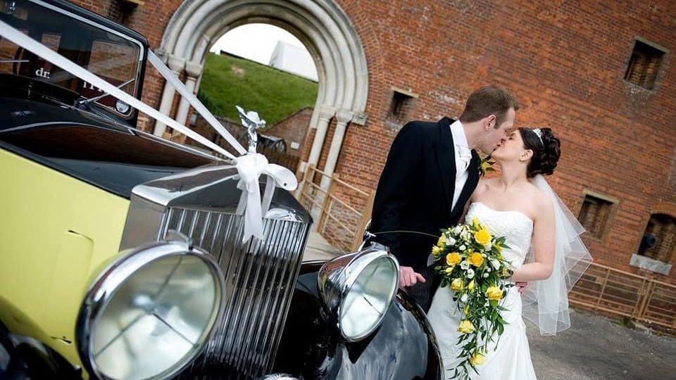 bride and groom kissing with vintage car in the foreground and Fort Nelson in the background