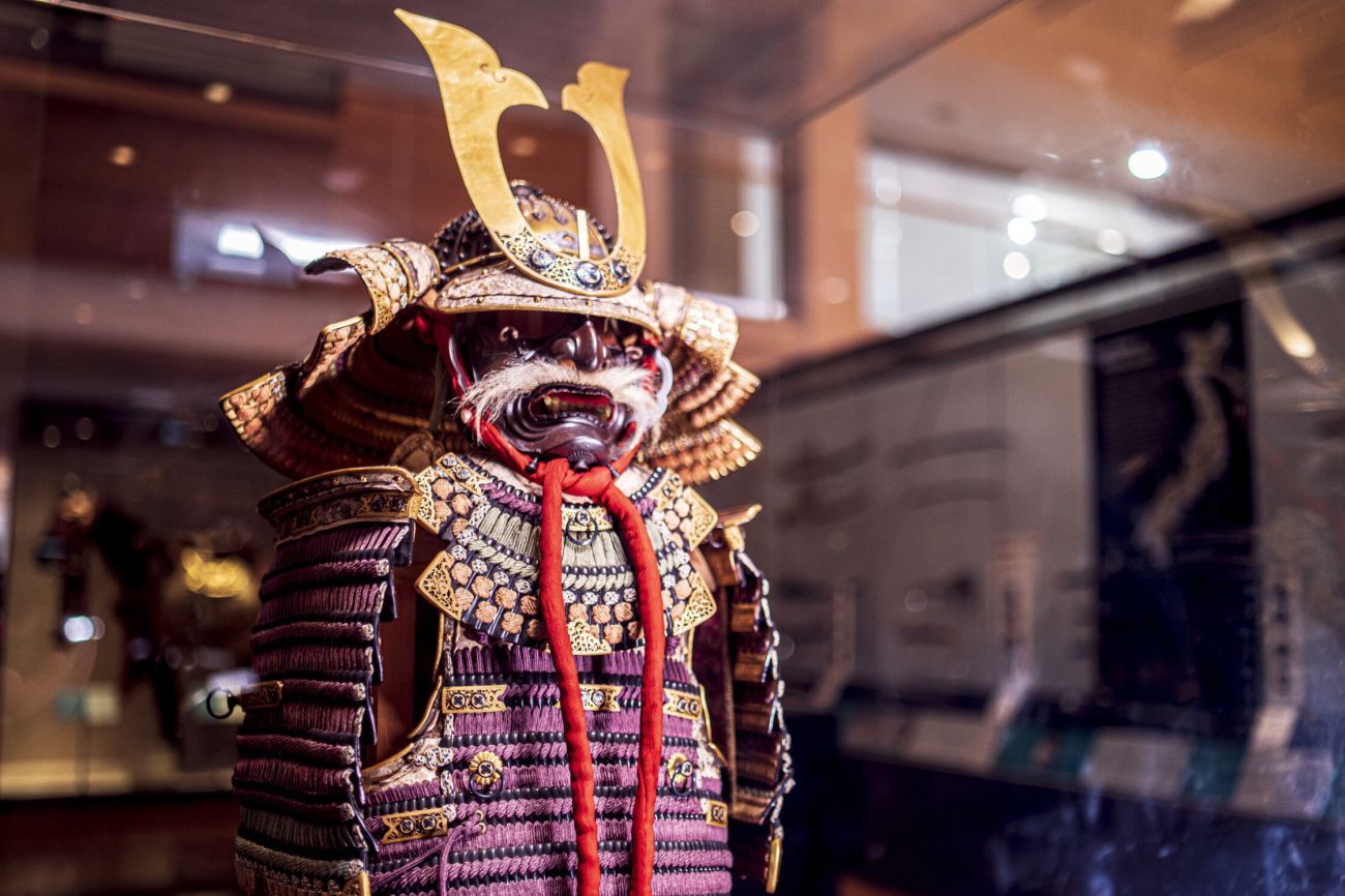 a purple and gold Samurai armour with a fearsome mask in the form of a demon with white whiskers stands guard in the gallery