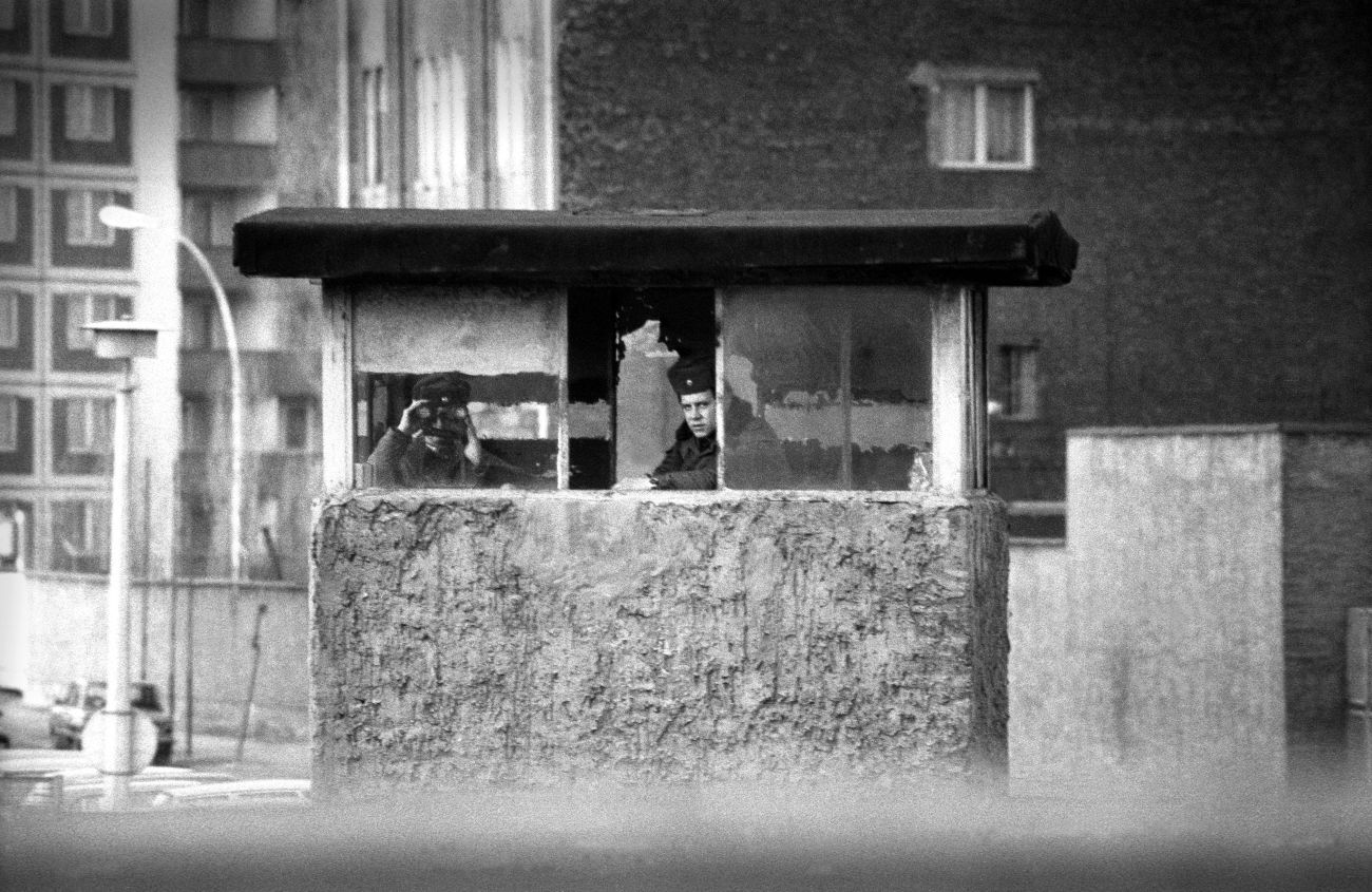 A black and white image showing two East German border guards sat in a concrete hut looking out of binoculars. They are surrounded by concrete plattenbau. 