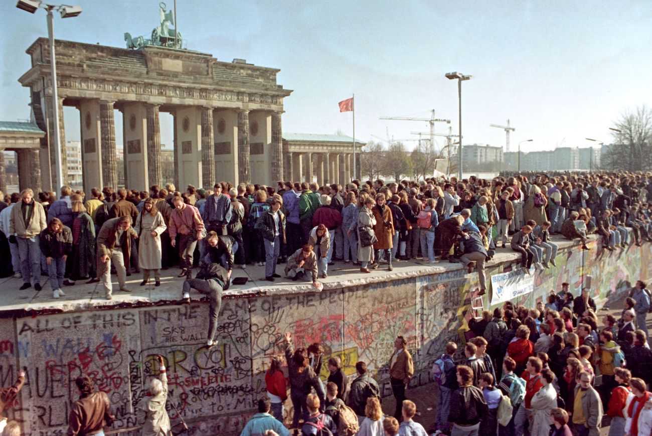 A large crowd of people are gathered at Berlin's Brandenburg Gate. Some are standing on top of the Berlin Wall