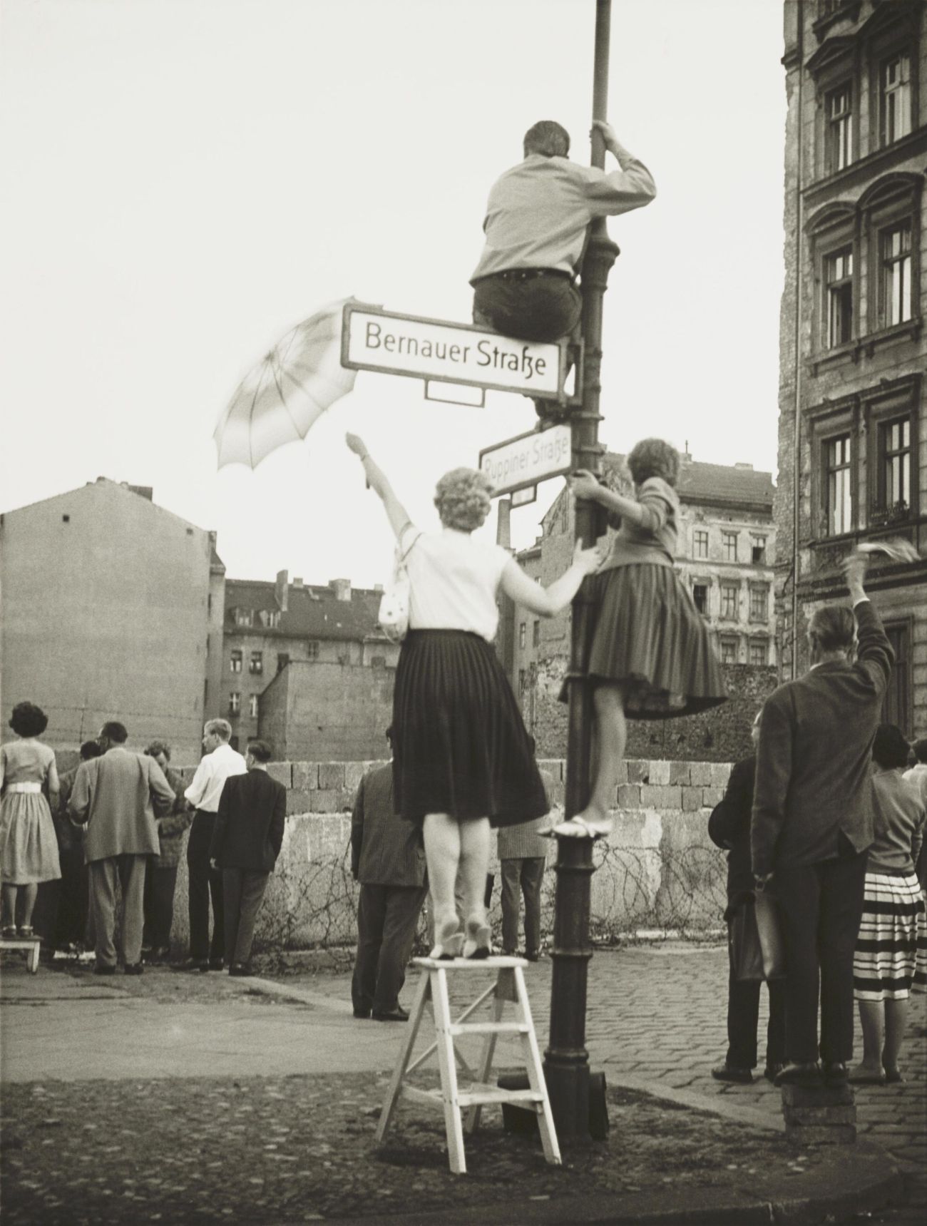 A black and white image showing West Berliners at the wall on Bernauer Strasse. There is a woman on a step ladder and two women have climbed a lamppost. Others are standing at the wall, looking across at their East Berlin friends and family