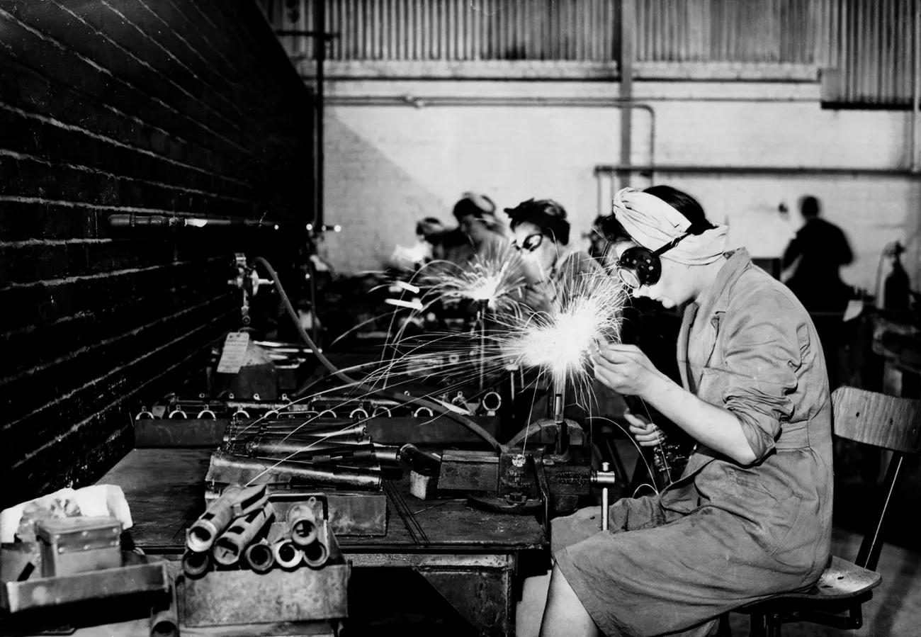 Women wearing coveralls, headscarves and goggles welding gun parts in a factory during Second World War