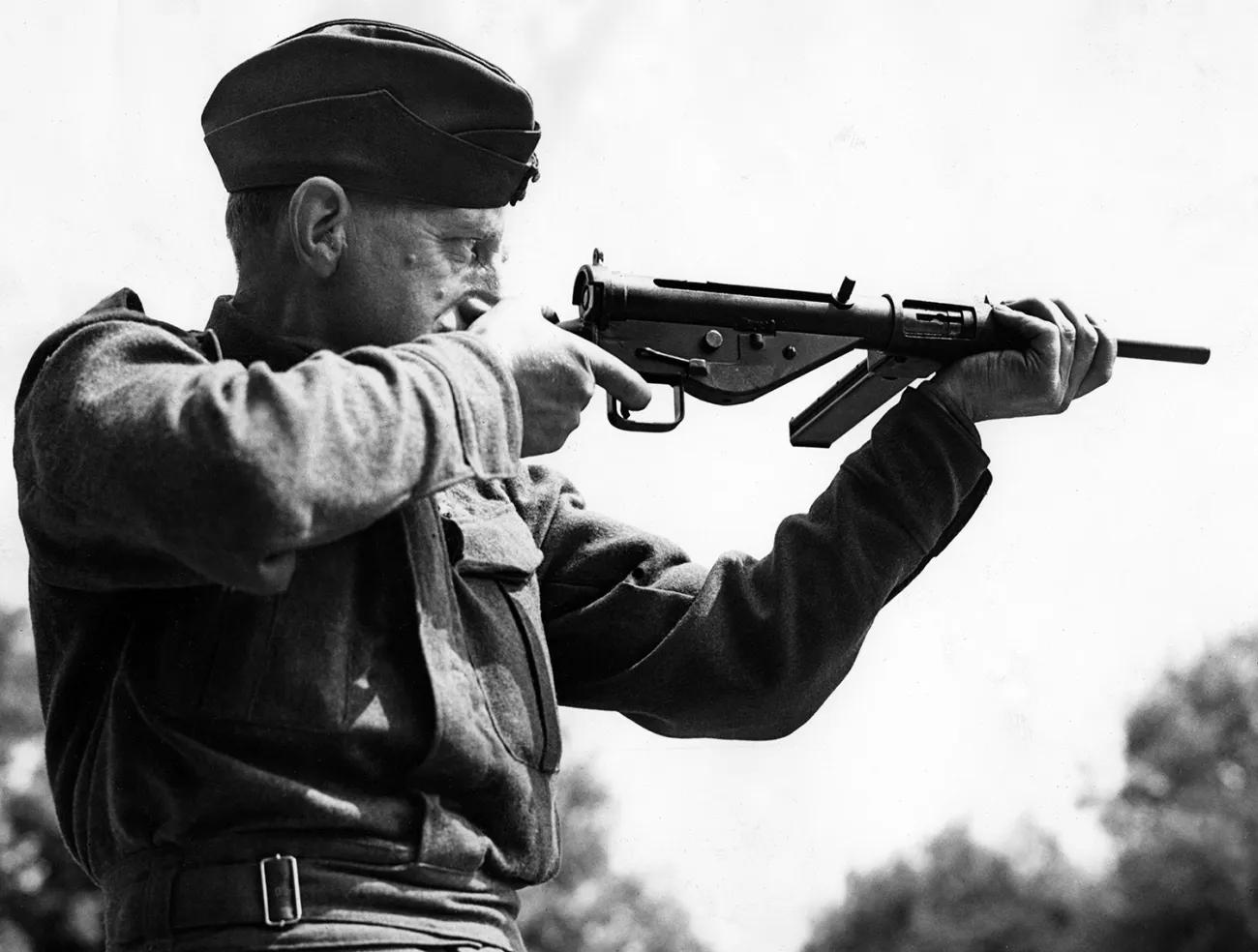 A man in second world war British army uniform firing a submachine gun