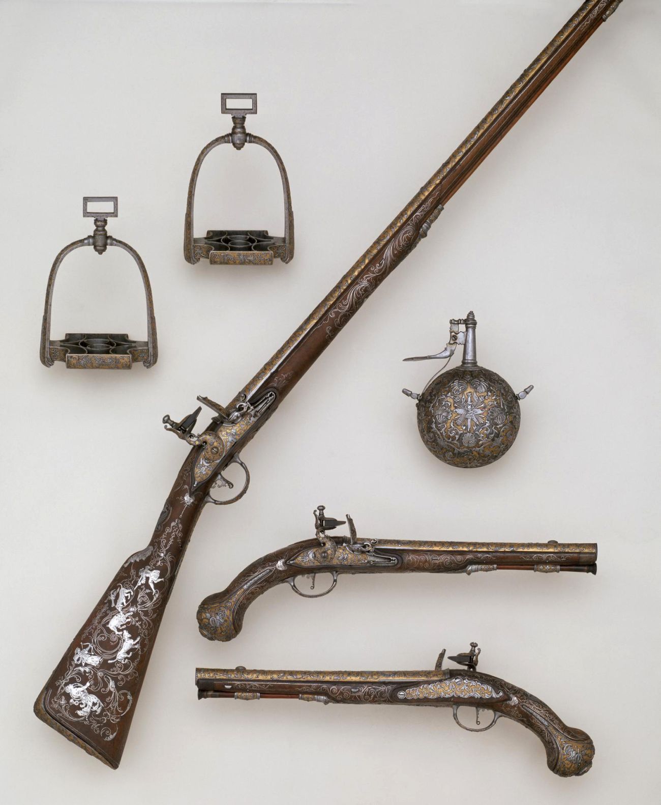 A set of similar highly decorated items including pair of stirrups, musket, pair pistols, powder flask