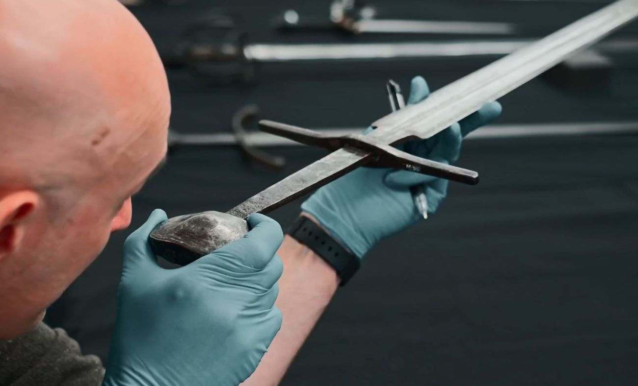 A swordsmith holds a sword up to his eye and carefully examines it along its length