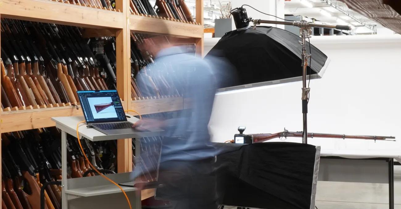 Photographer looking at image on a laptop with firearm on a table with photography lights and equipment around it.