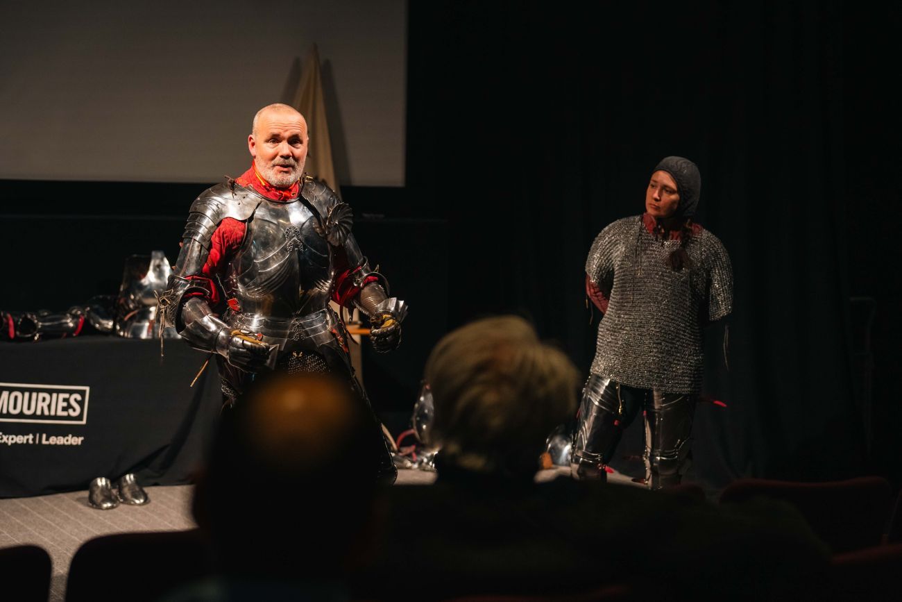Man in armour giving a demonstration to the audience with female wearing chainmail beside him.