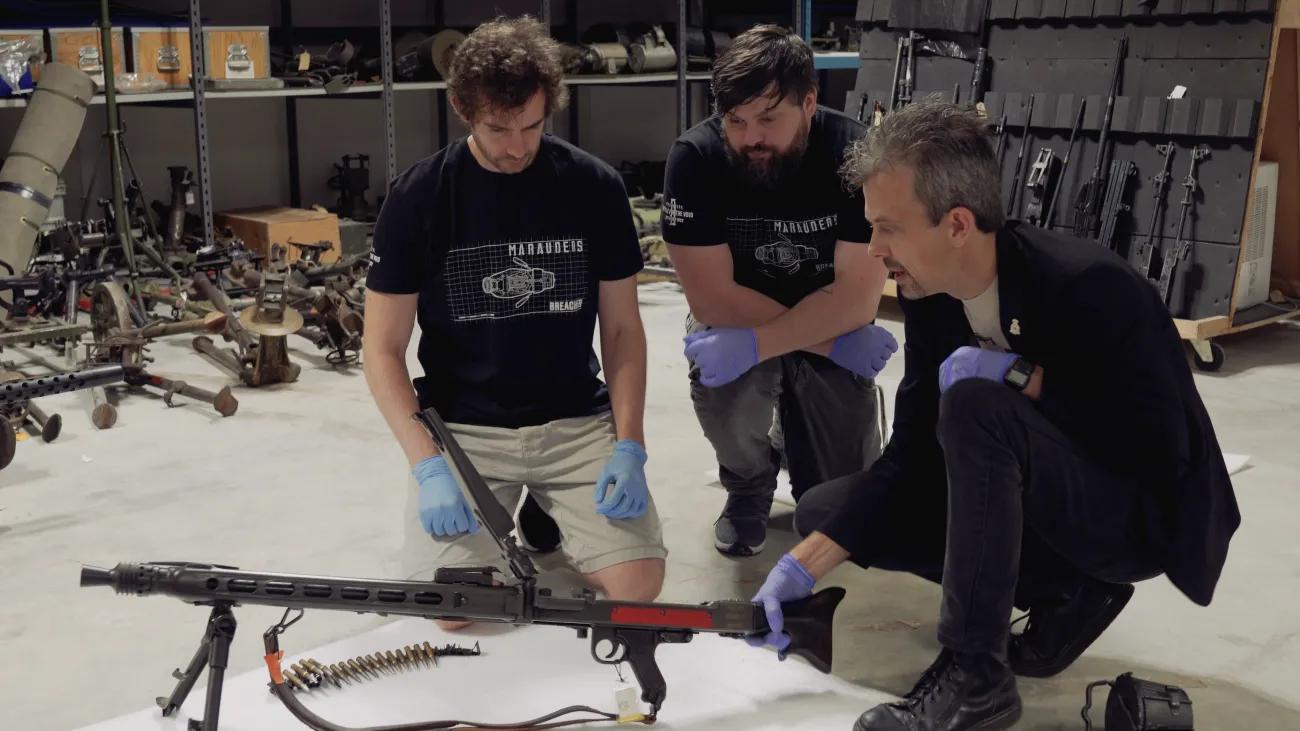 Jonathan Ferguson talking to two visitors whilst holding firearm and also other firearms in the background