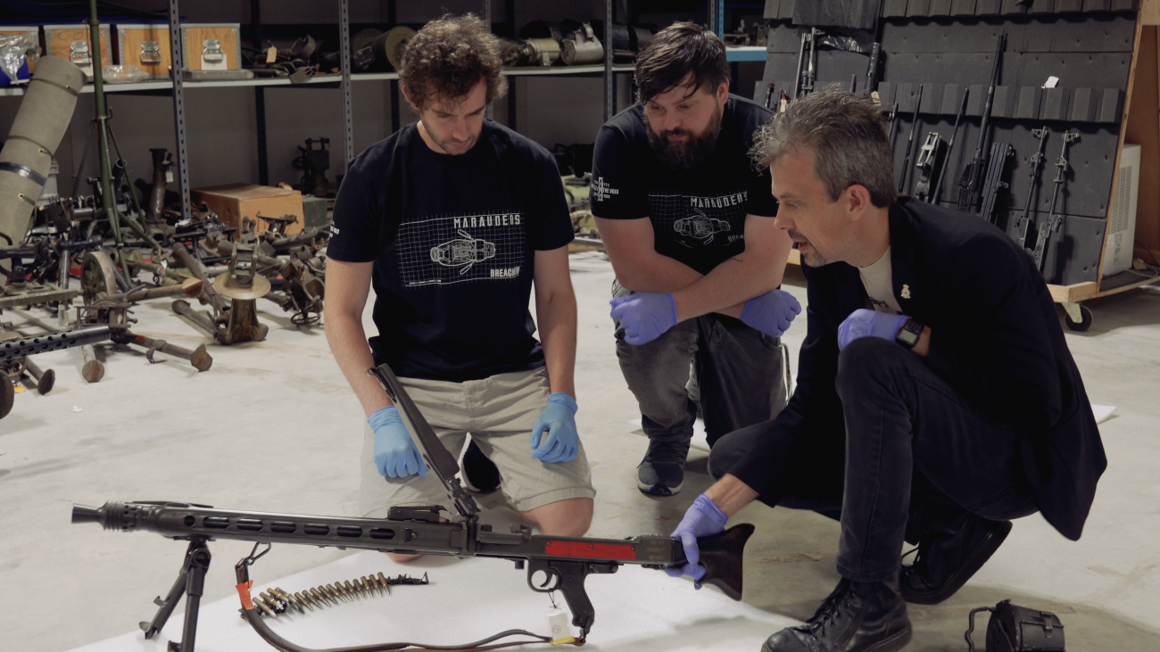 Jonathan Ferguson talking to two visitors whilst holding firearm and also other firearms in the background