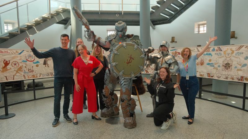 Project team members pose with Doom character in front of slayeux tapestry in the hall of steel