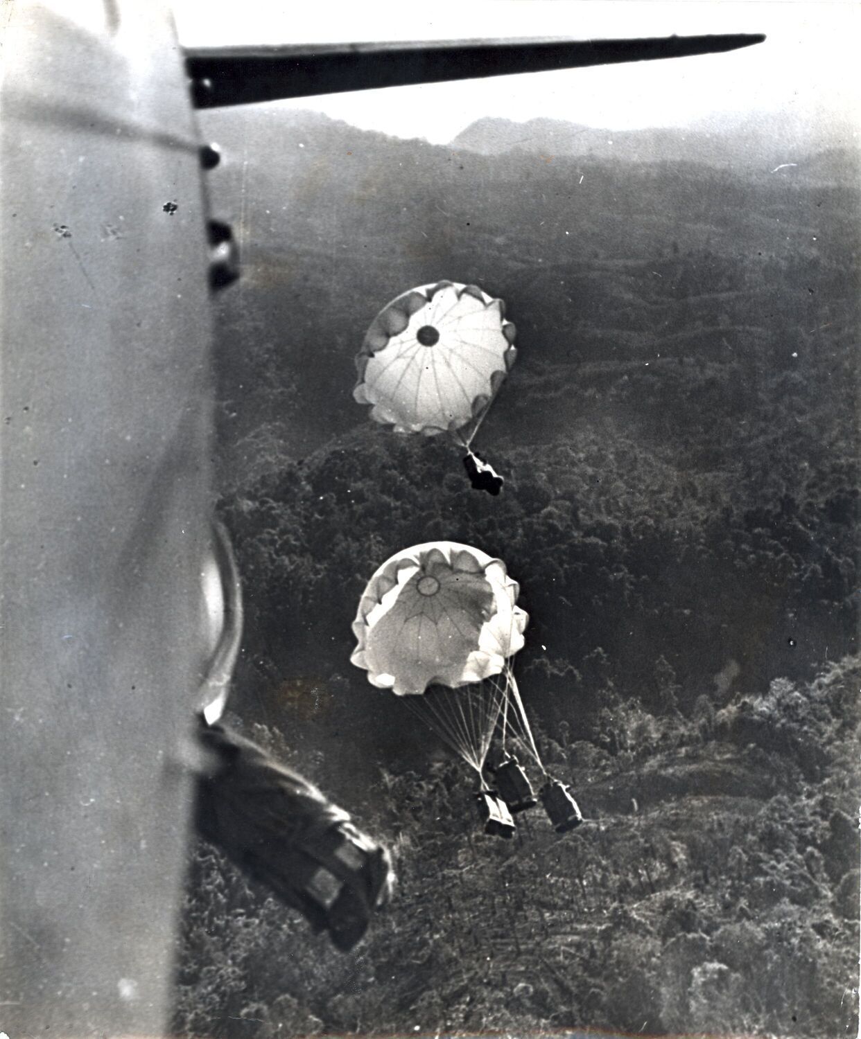 A black and white photo taken from the plane of supplies being airdropped to soldiers waiting in a jungle