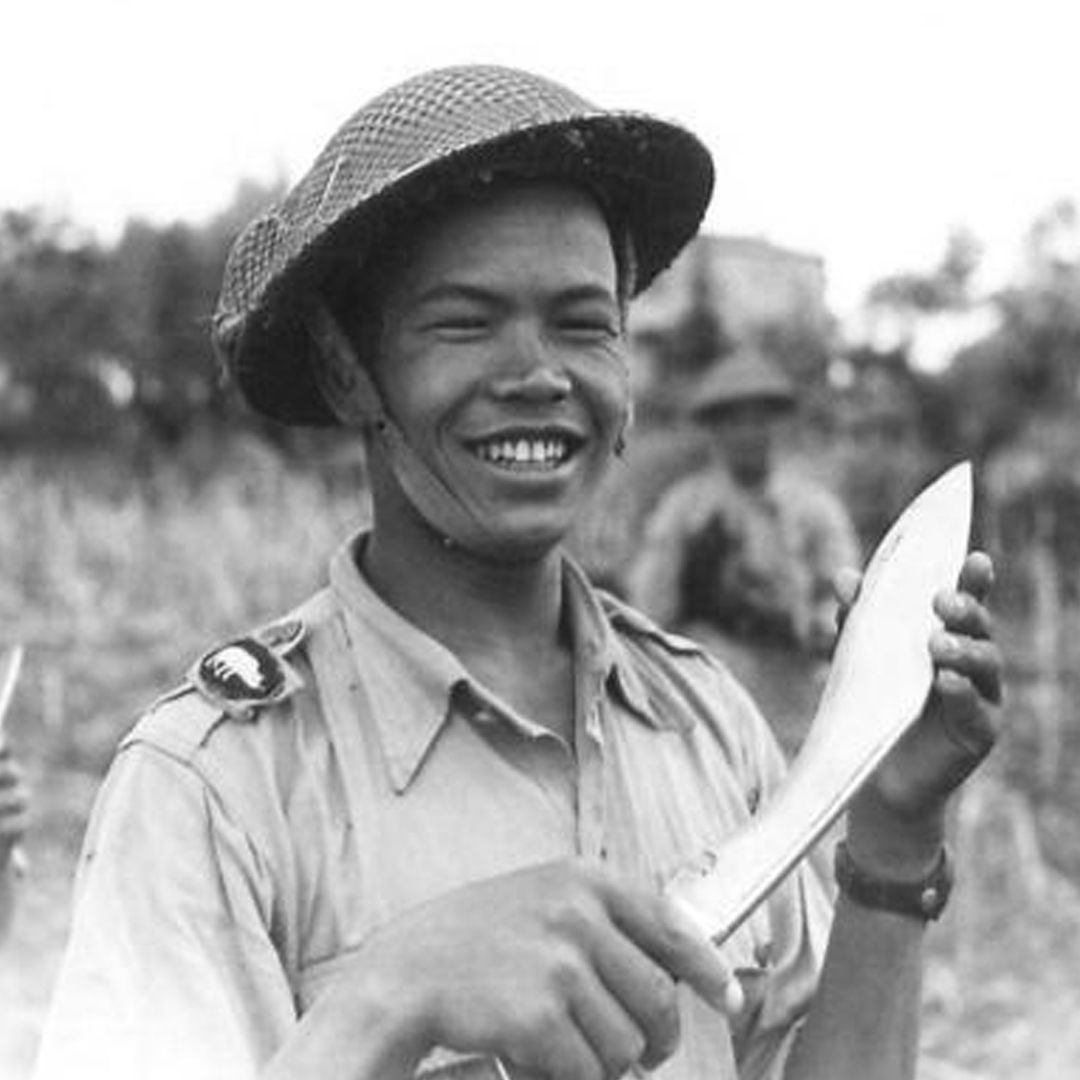 A smiling soldier in khaki uniform and metal helmet poses with a kukri knife