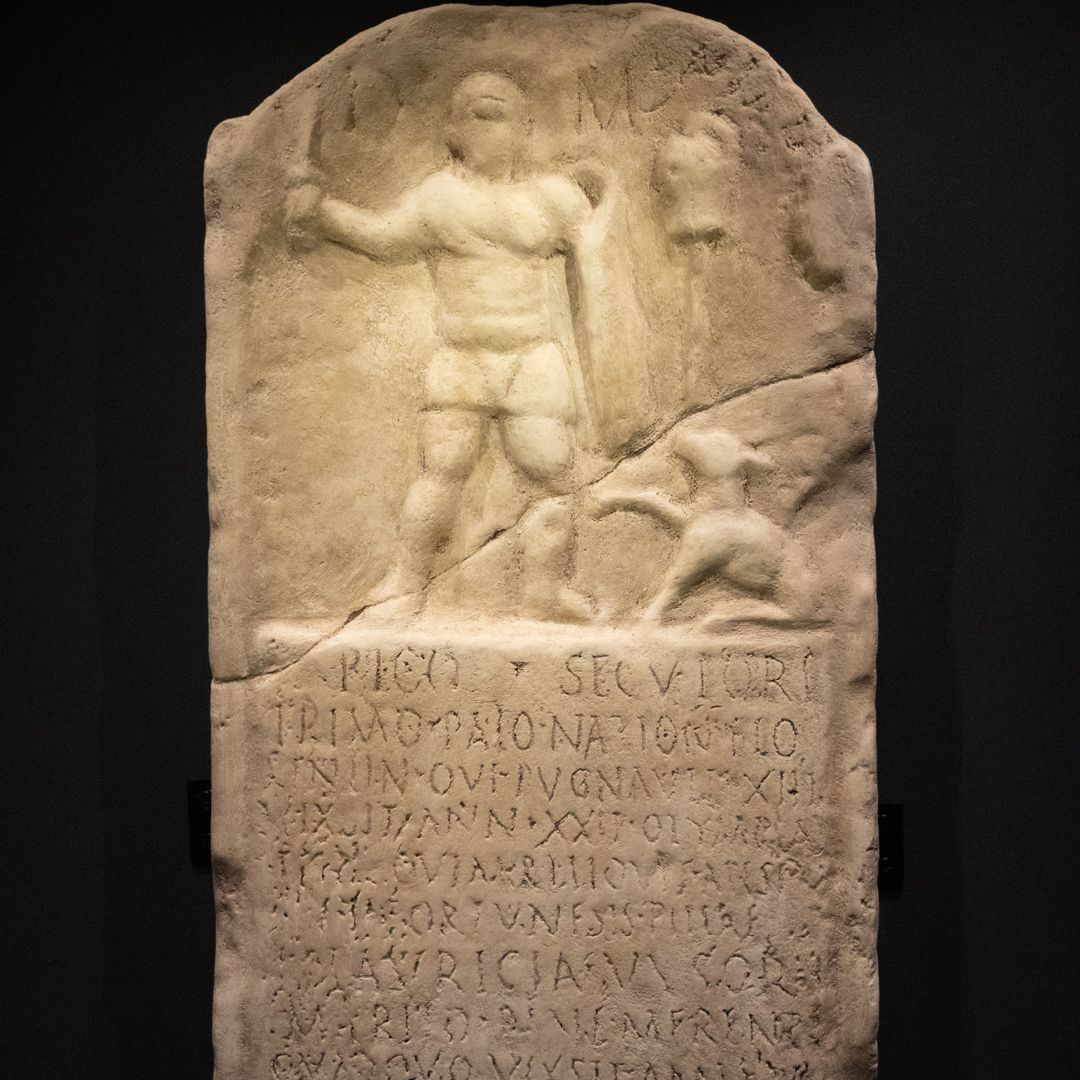 A sandy coloured stone tablet depicting a gladiator and his dog, with his life story inscribed below
