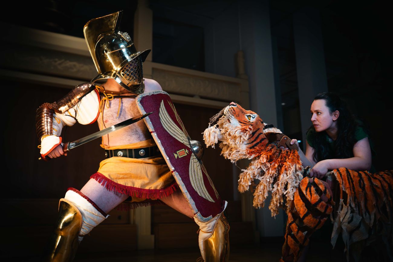 Gladiator in traditional armour with sword and shield confronts a large tiger puppet operated by a person