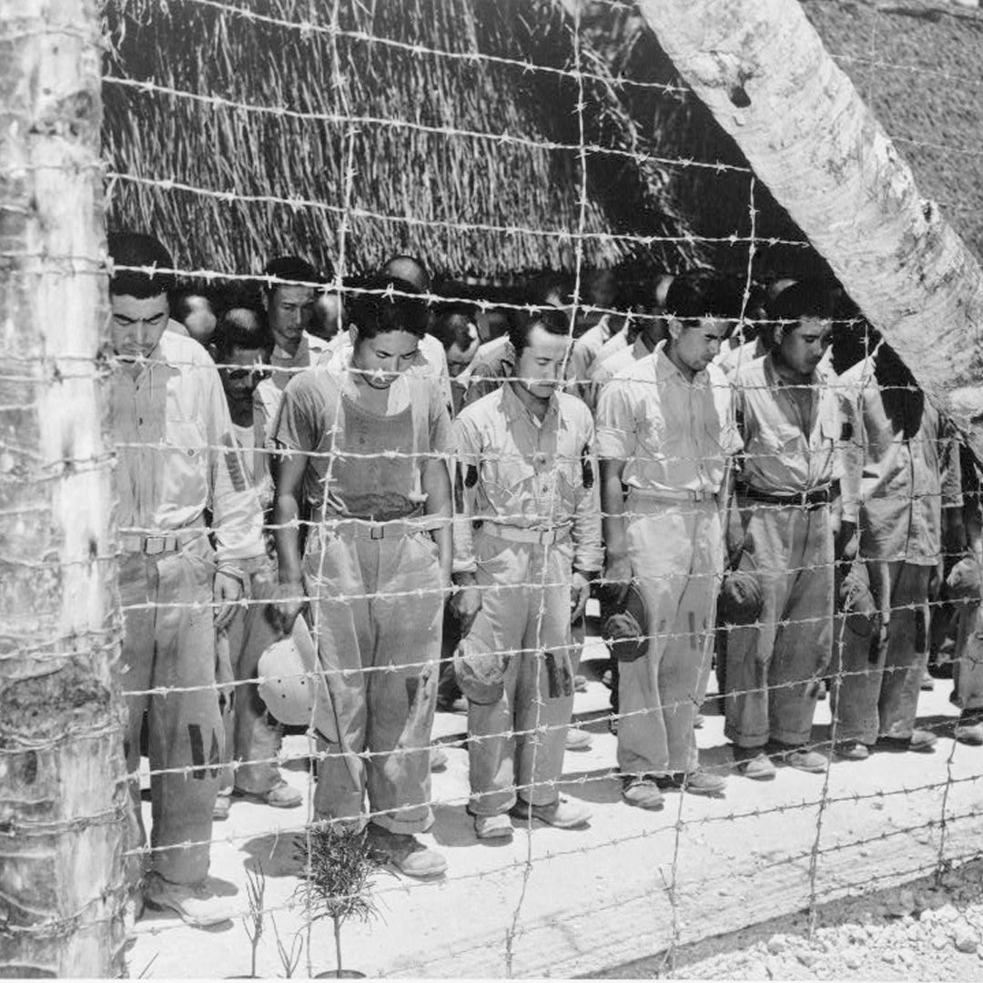 A black and white photograph of Japanese prisoners of war bowing their heads from behind a barbed wire fence.